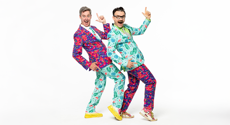 Two men in brightly coloured suits point their index fingers in the air and smile. 