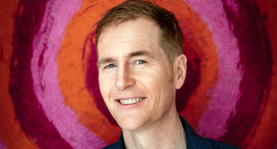 A man smiles at the camera, with pink and red circles behind him.