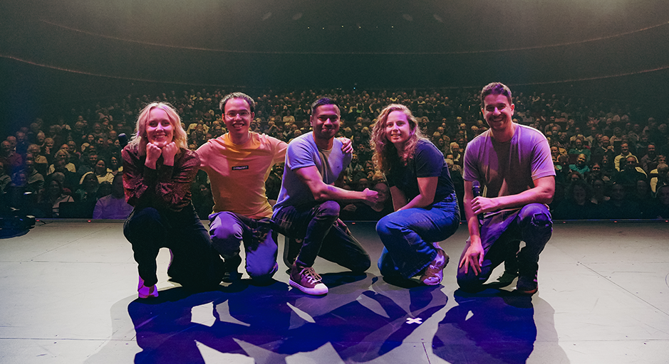 A photo of a group of comedians on stage, behind them is a sold-out audience.