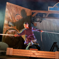 A masculine person in traditional Chinese clothing holds two ropes together in thick stage fog.