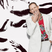 Todd McKenney wears a white suit with a floral red shirt, against and black and white print background.