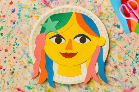 Colourful paper craft face with multicoloured hair and a green star, arranged on a paint-splattered background, with scissors and coloured paper to the side.
